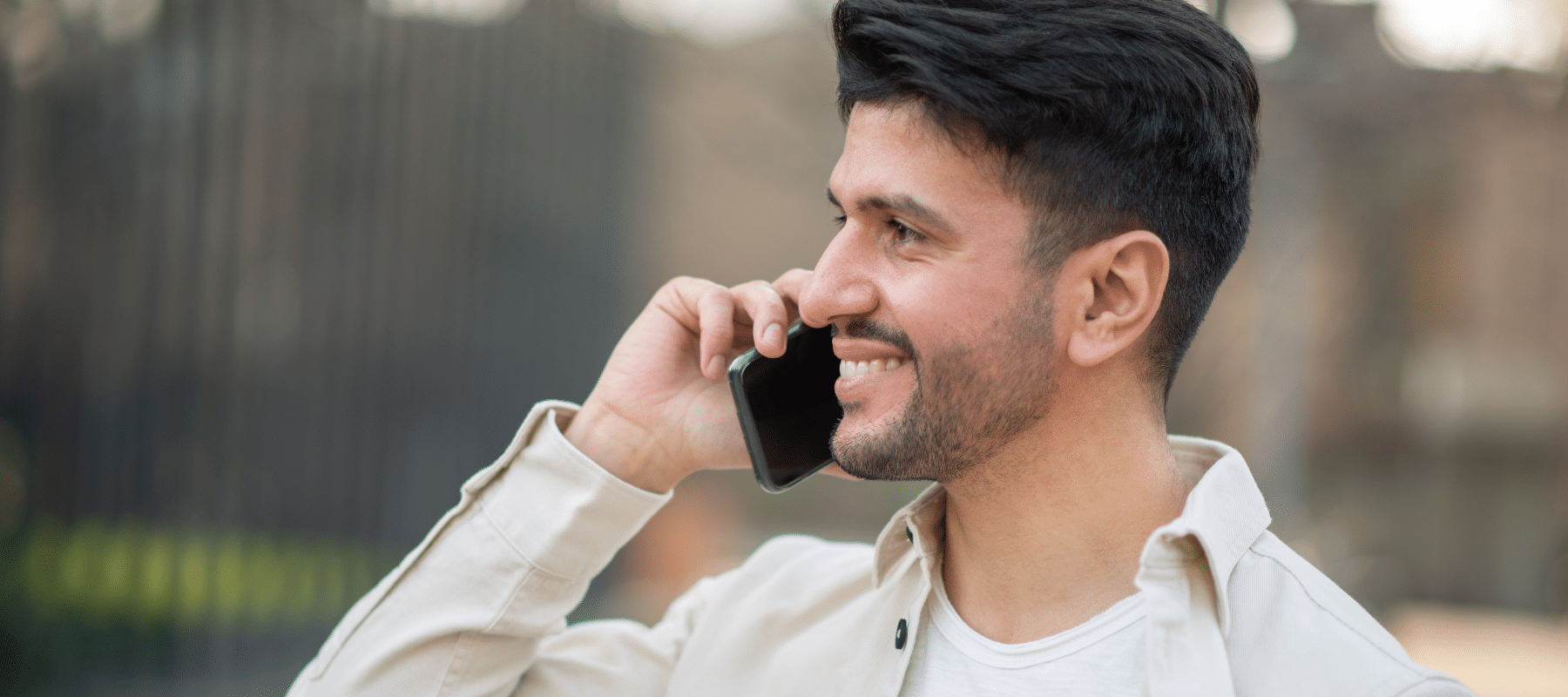 A smiling man talking on his smartphone outdoors, wearing a light-colored jacket.