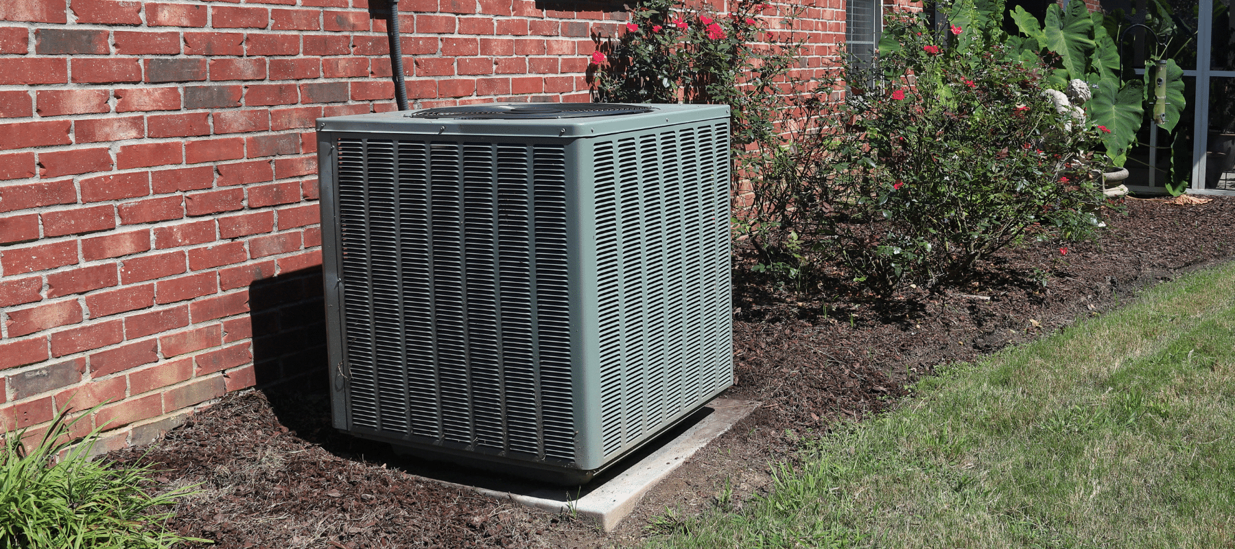 Outdoor air conditioning unit next to a brick house surrounded by mulch and landscaping